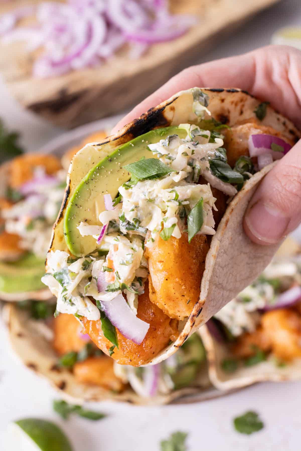 A hand lifts a single Baja shrimp taco from a plate.