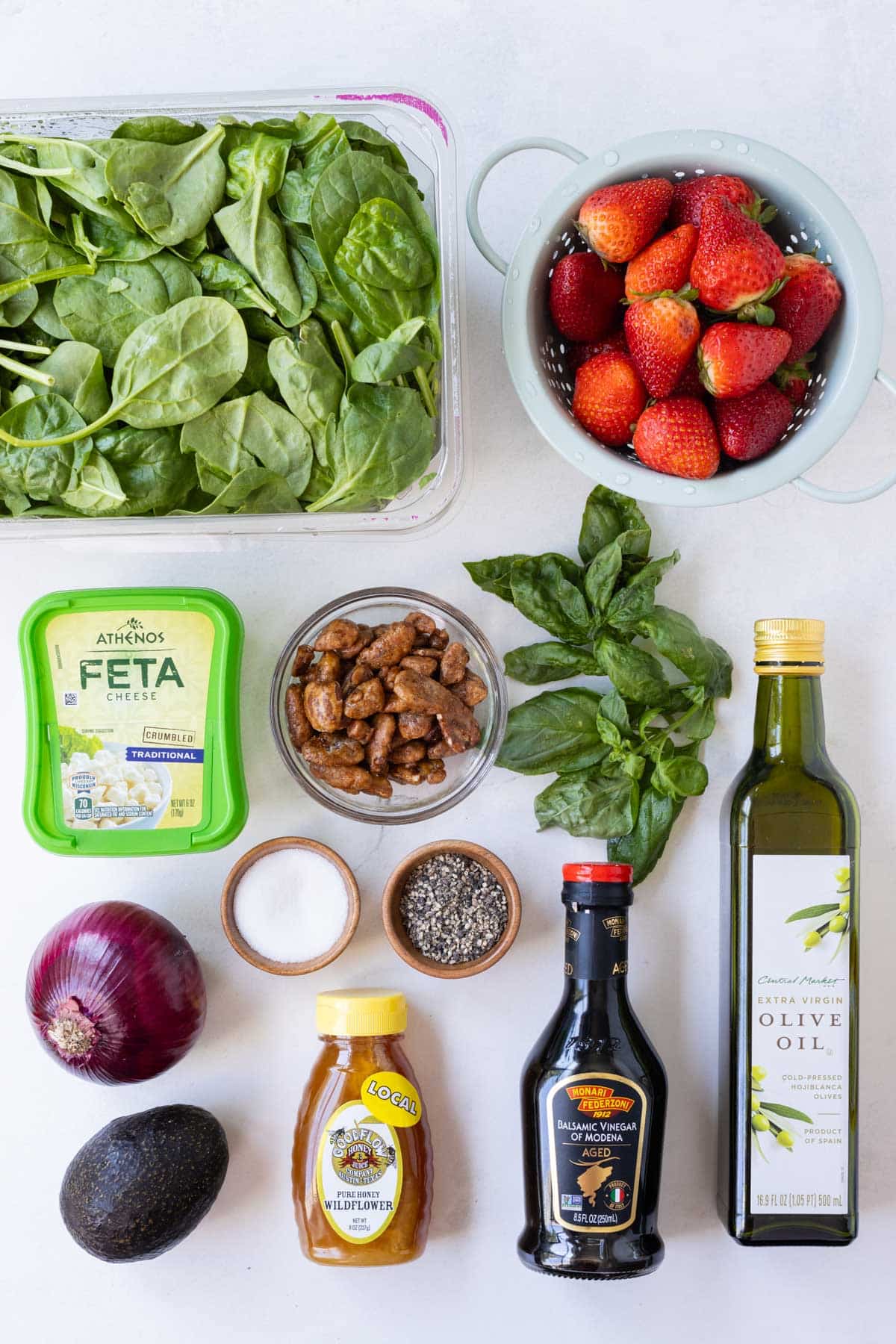Spinach, strawberries, feta, candied pegans, basil, red onion, avocado, honey, balsamic vinegar, olive oil and salt and pepper are arranged as ingredients for a salad.