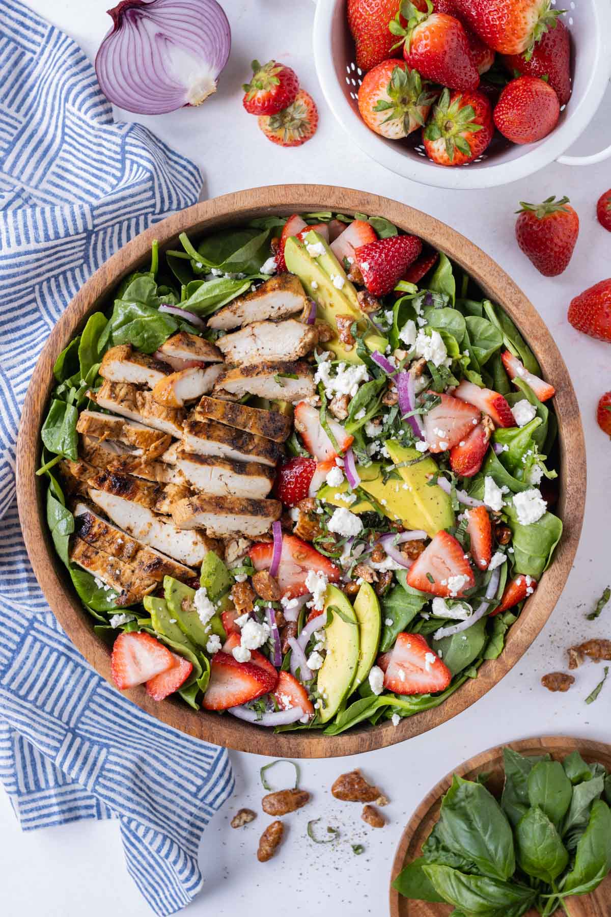 A bowl of strawberry spinach salad with balsamic chicken next to a blue dish towel, a bowl of strawberries, and fresh basil. 