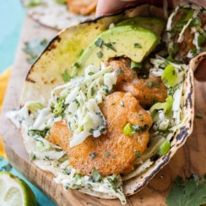 A hand is picking up a Baja Shrimp Taco with a fresh and creamy slaw and avocado slices.