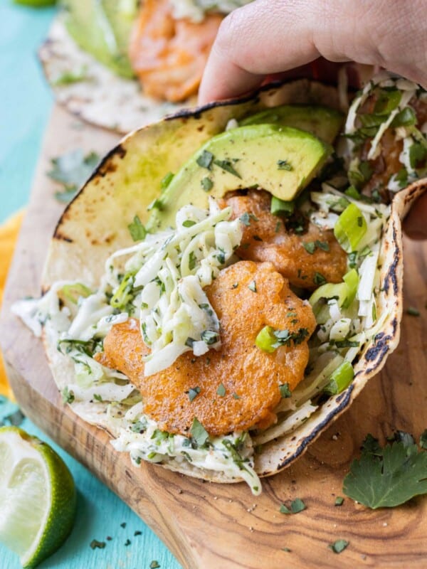 A hand is picking up a Baja Shrimp Taco with a fresh and creamy slaw and avocado slices.