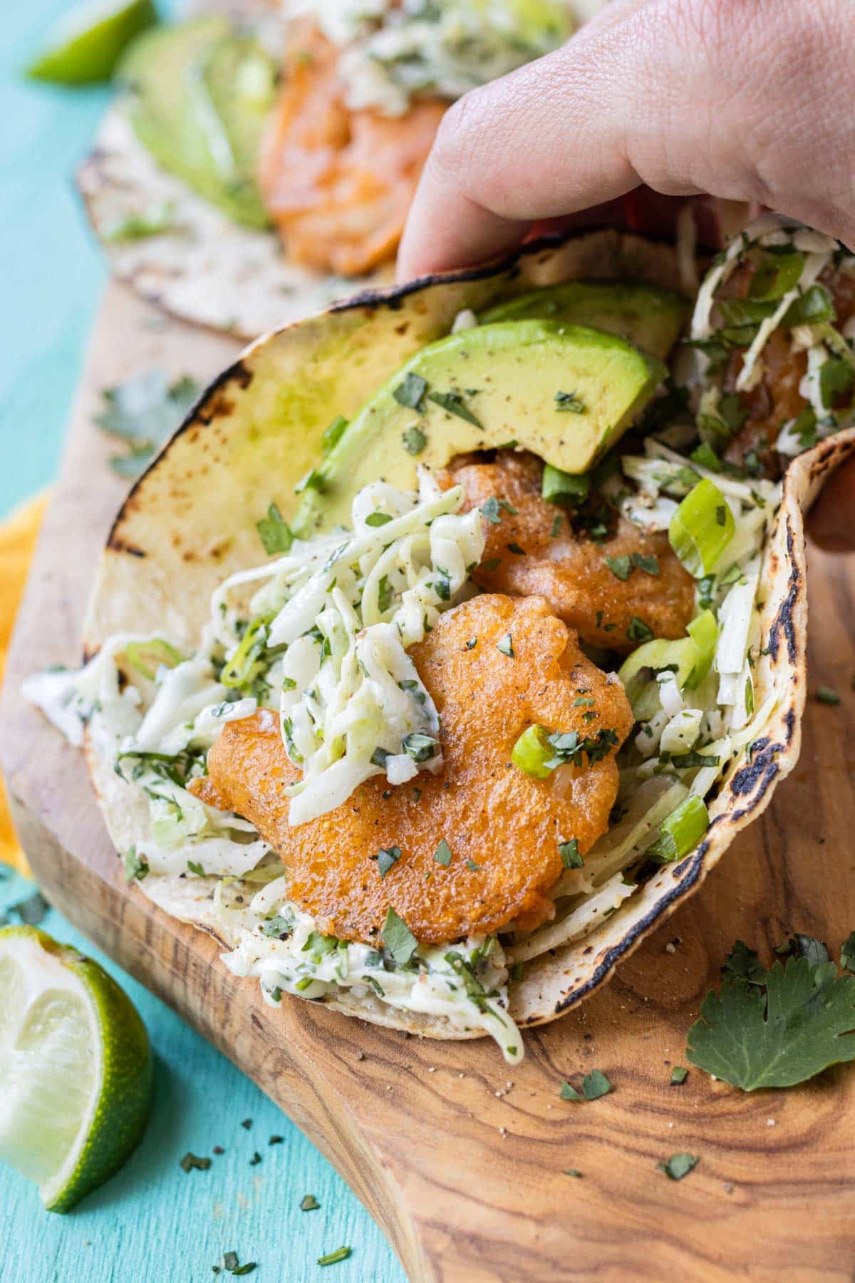 A hand is picking up a Baja Shrimp Taco with a fresh and creamy slaw and avocado slices.