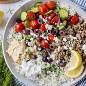 Greek turkey, cucumber-tomato salad, and other toppings are served on a bed of rice with a lemon wedge.