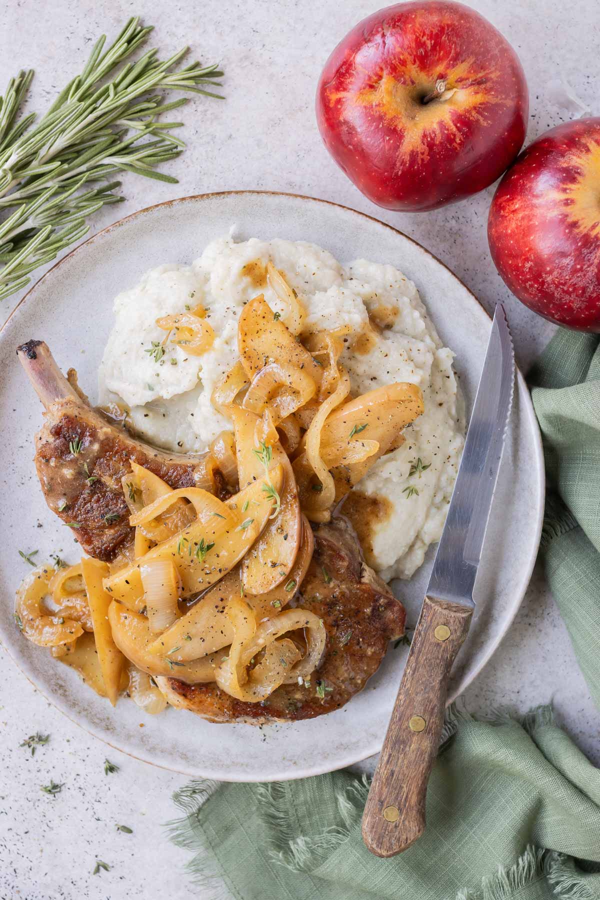 Bone in pork chops, apples, and onions are served on a white plate with mashed potatoes.