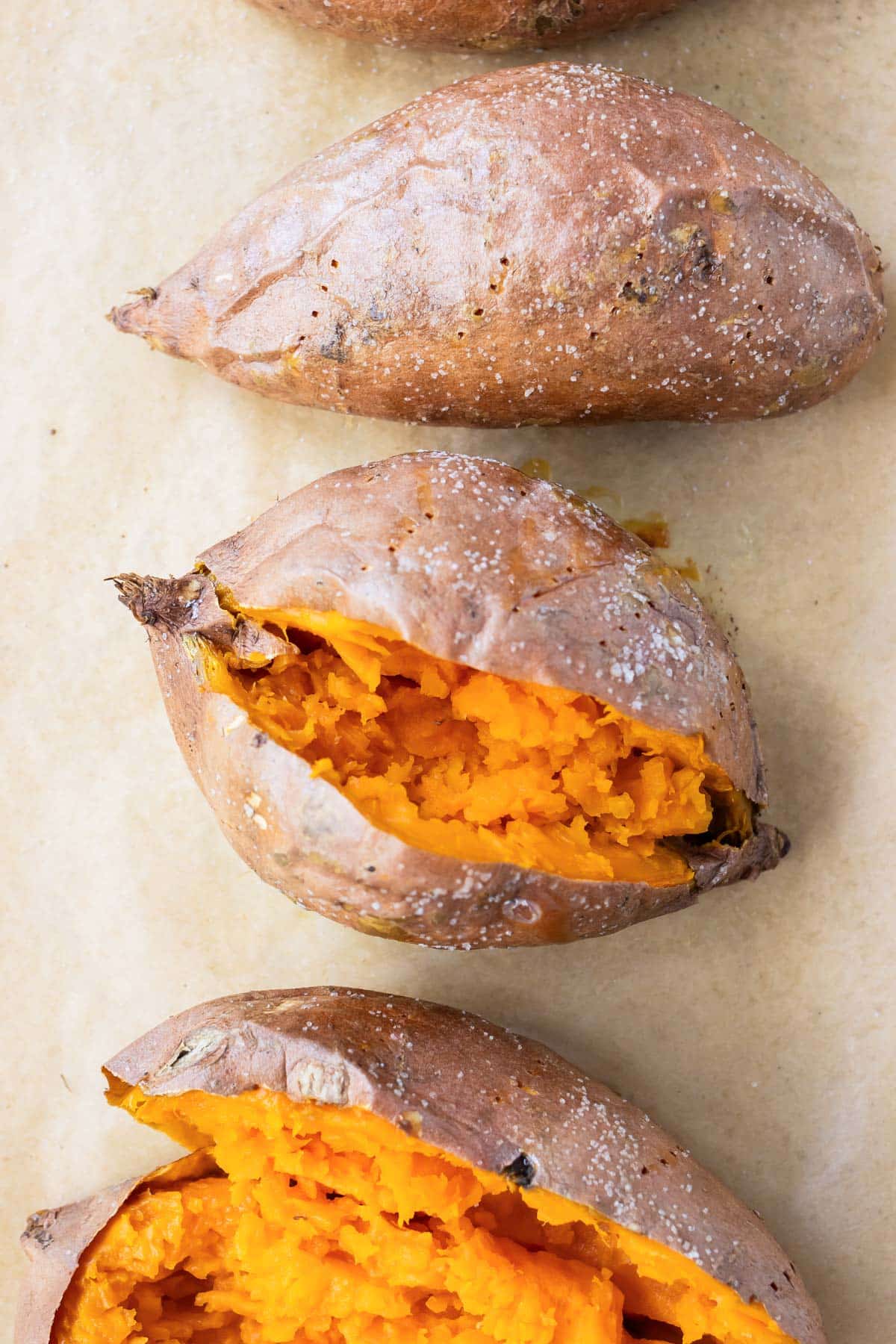 Baked sweet potatoes on a parchment paper lined baking sheet that are cut open with crispy skin.