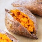 Four baked sweet potatoes on a baking sheet with crispy skin and fluffy insides.