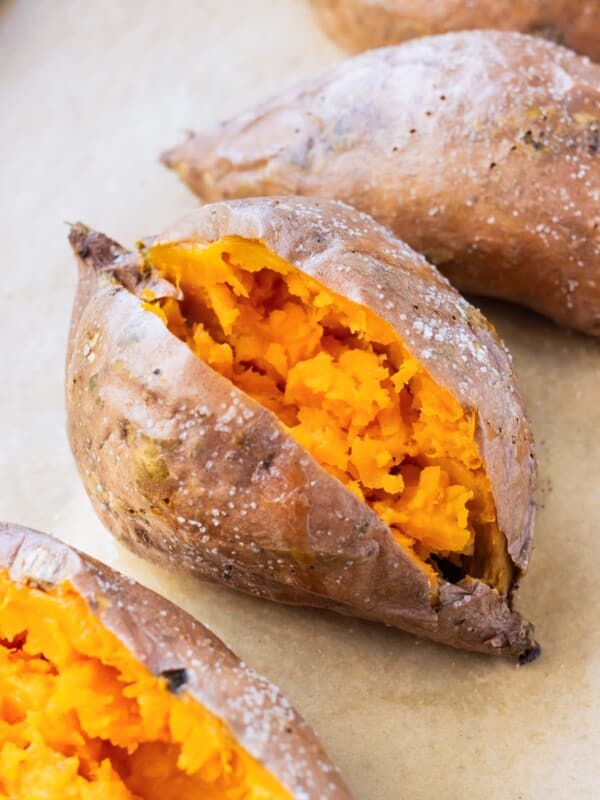 Four baked sweet potatoes on a baking sheet with crispy skin and fluffy insides.