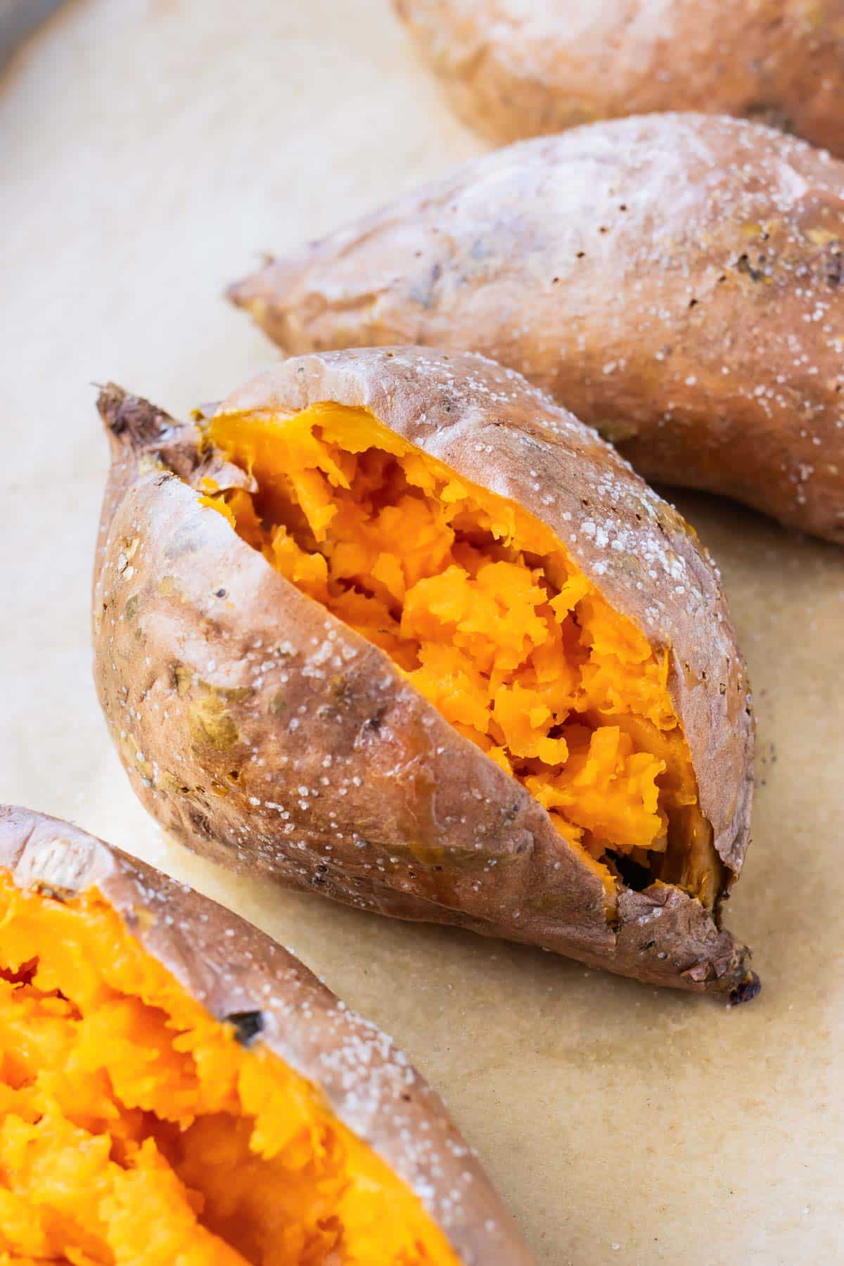 Four baked sweet potatoes on a baking sheet with crispy skin and fluffy insides.