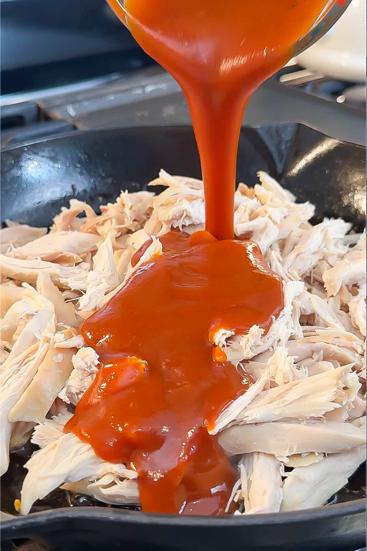 BBQ sauce being poured into a skillet with shredded chicken.