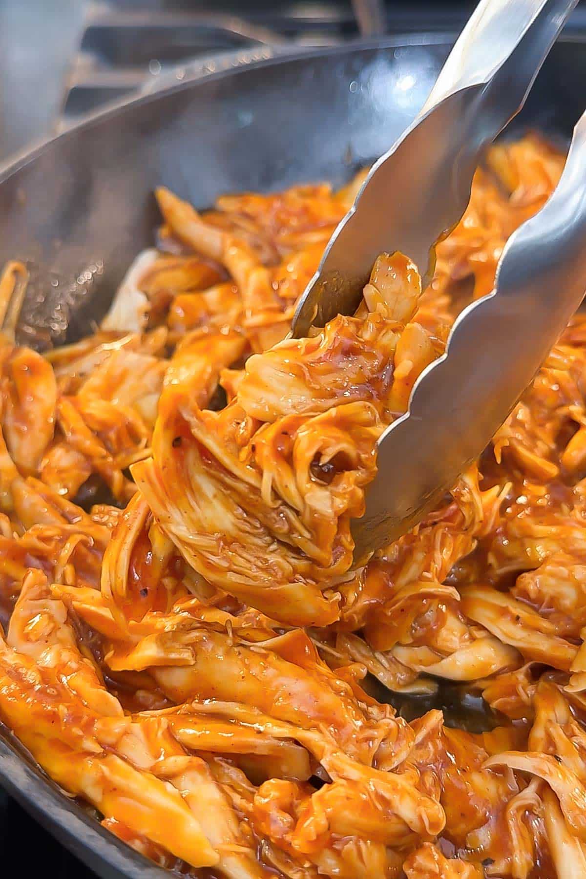 Shredded chicken being tossed with tongs in a skillet.