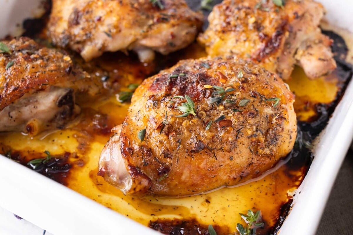 A close-up picture of a bone-in chicken thigh in a baking dish with a honey mustard sauce.