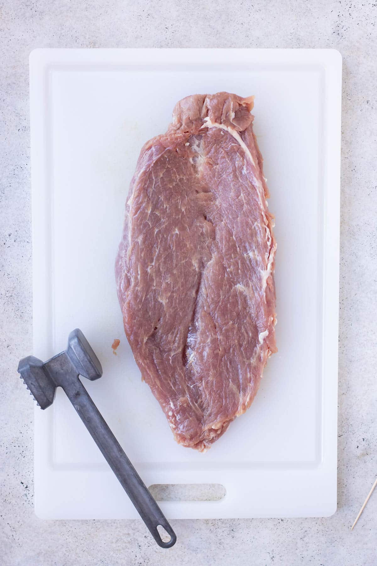 Pork tenderloin split open, on a plastic cutting board with a meat mallet.