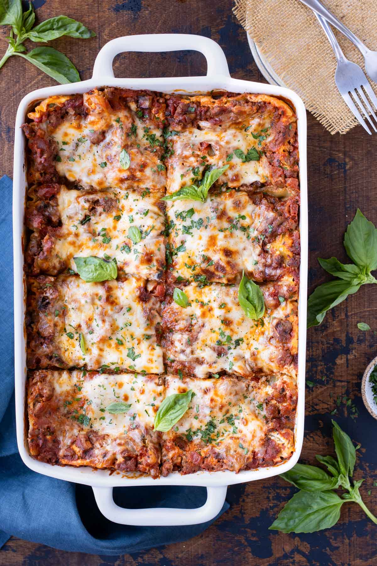 A lasagna recipe is in a white baking dish with basil leaves on top next to dinner plates.