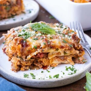 A piece of an easy lasagna recipe is on a white plate with a fork next to it and parsley sprinkled around.