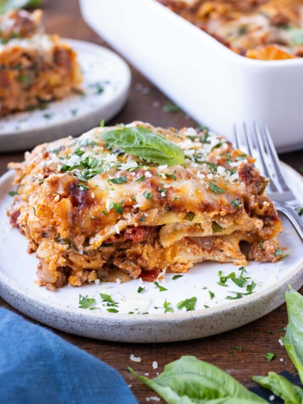 A piece of an easy lasagna recipe is on a white plate with a fork next to it and parsley sprinkled around.