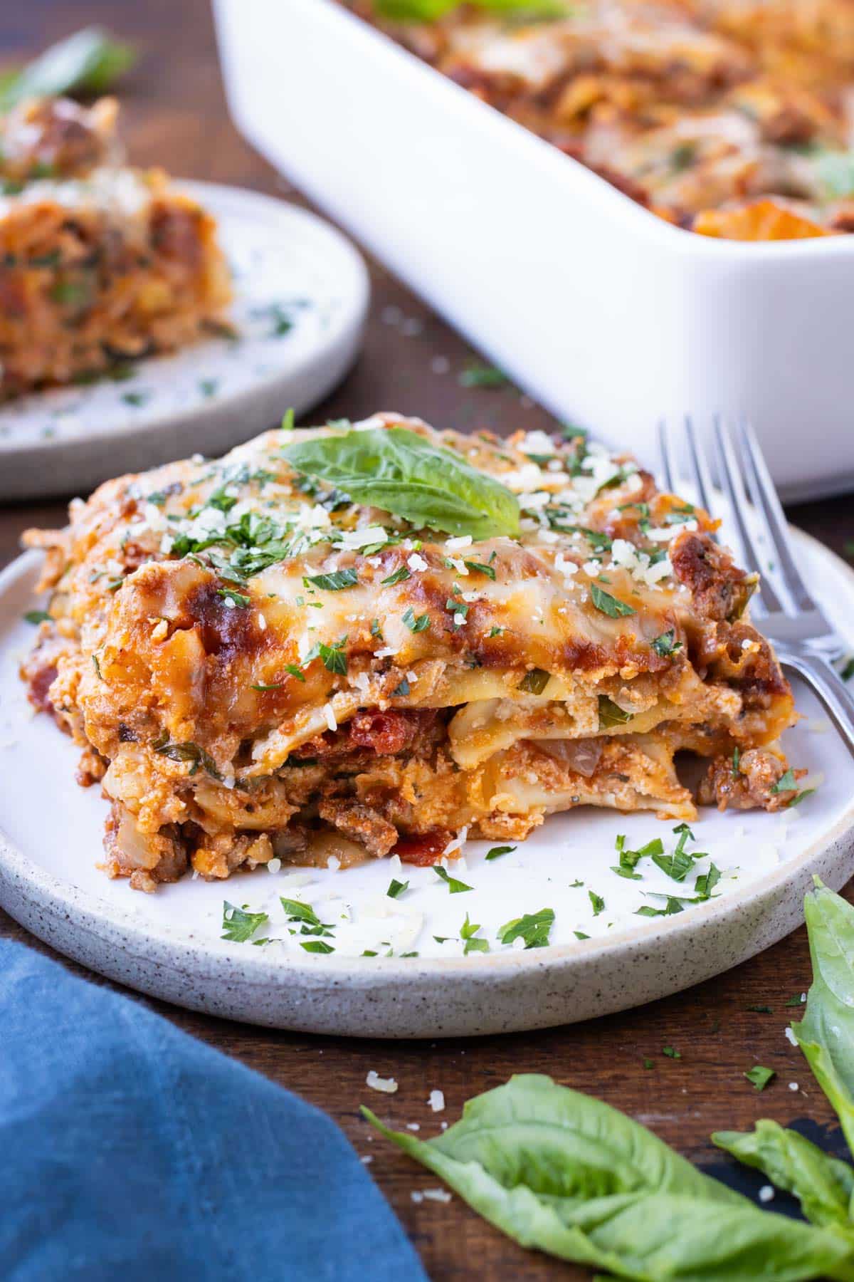 A piece of an easy lasagna recipe is on a white plate with a fork next to it and parsley sprinkled around.