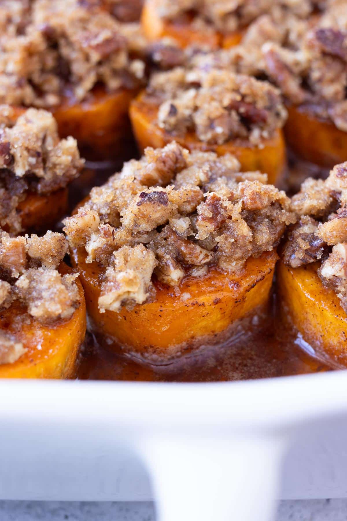 A dish full of melting sweet potatoes, topped with a pecan crumble and maple sauce.