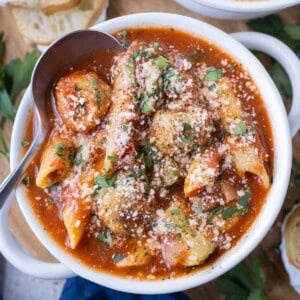 A white bowl full of Chicken Parmesan Meatball Soup with garlic butter cheese sandwiches surrounding it.