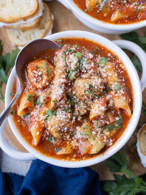 A white bowl full of Chicken Parmesan Meatball Soup with garlic butter cheese sandwiches surrounding it.