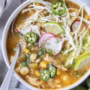 Chicken pozole verde with hominy is in a white bowl with a spoon and topped with cabbage, radishes, and avocado.