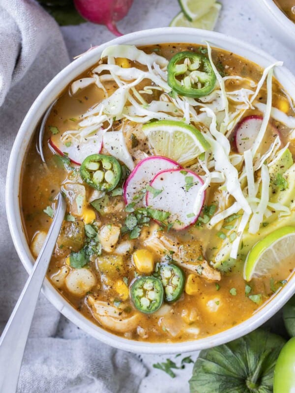 Chicken pozole verde with hominy is in a white bowl with a spoon and topped with cabbage, radishes, and avocado.