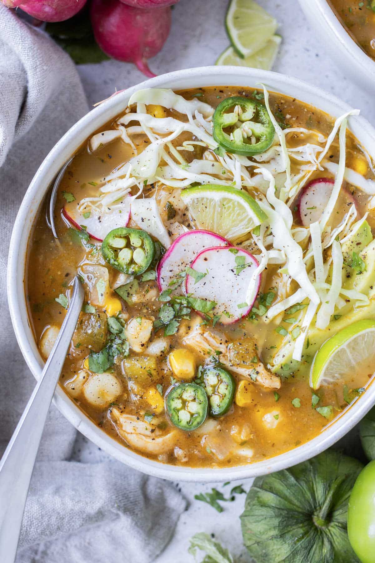 Chicken pozole verde with hominy is in a white bowl with a spoon and topped with cabbage, radishes, and avocado.