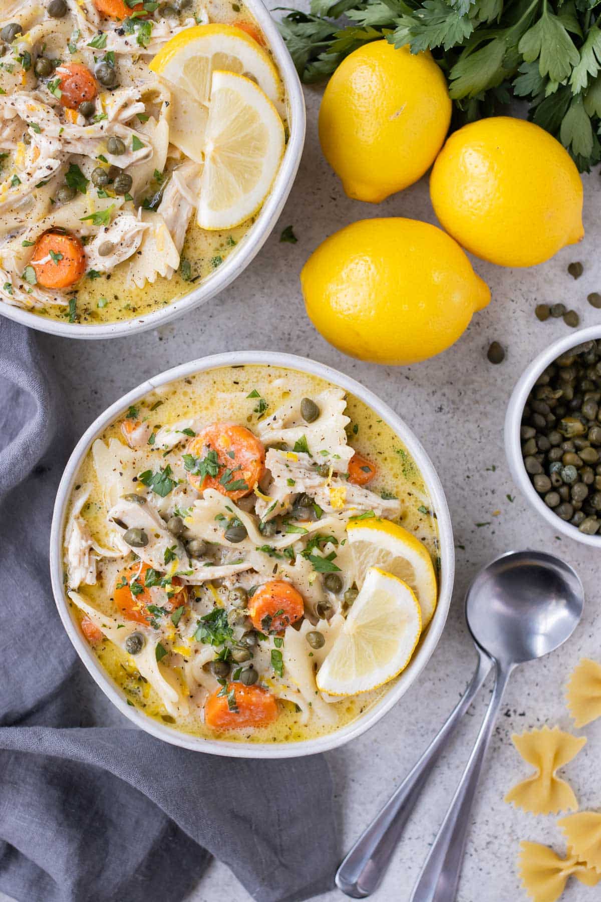 Two bowls of Lemon Chicken Piccata Soup are served next to lemons and capers.