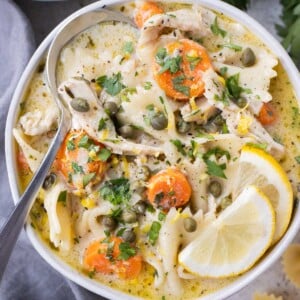 A bowl full of a creamy lemon chicken piccata soup with carrots, pasta, capers, and lemon slices.