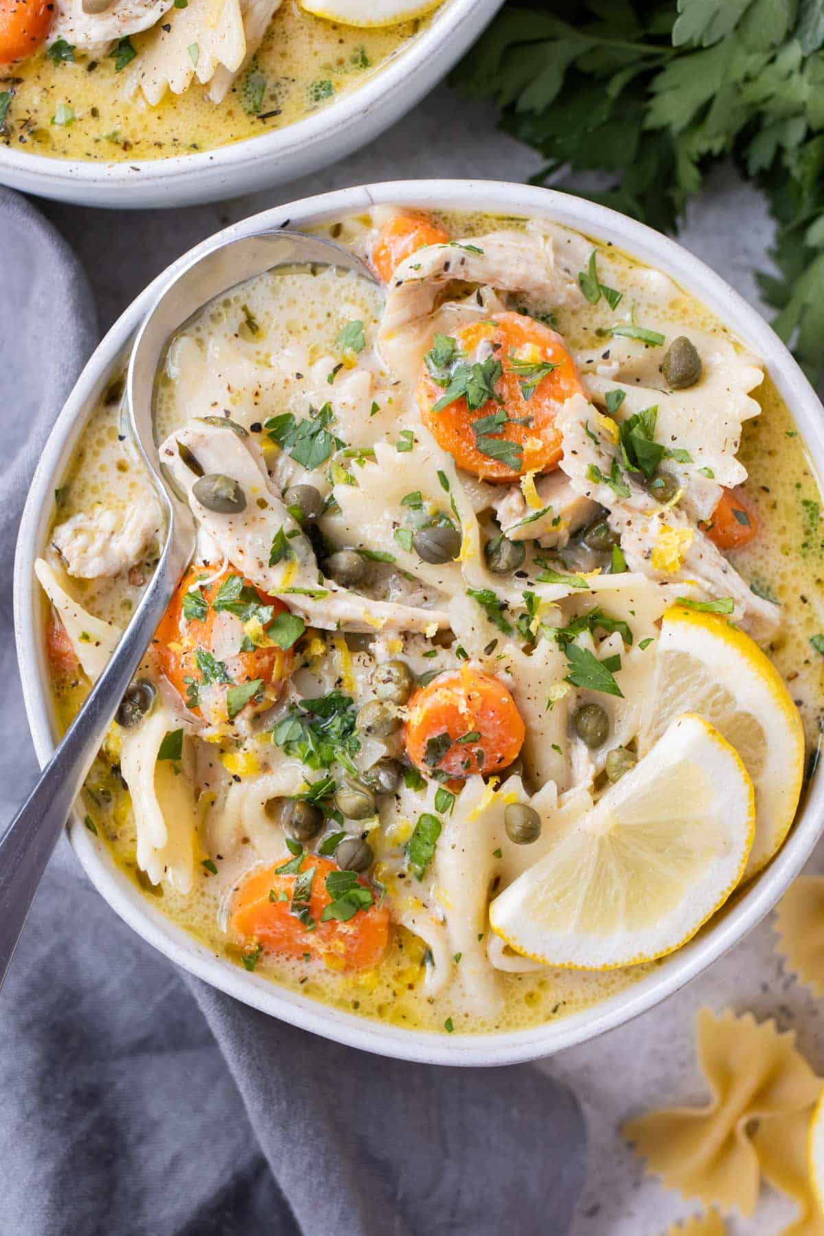 A bowl full of a creamy lemon chicken piccata soup with carrots, pasta, capers, and lemon slices.