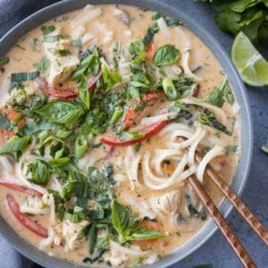 Chopsticks going into a bowl of Thai Red Curry Chicken Soup with fresh herbs and limes next to it.