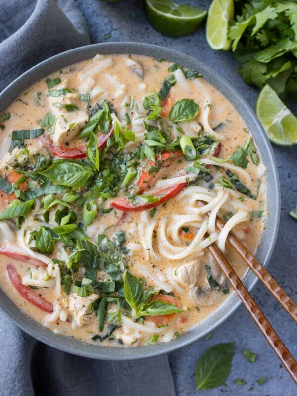 Chopsticks going into a bowl of Thai Red Curry Chicken Soup with fresh herbs and limes next to it.