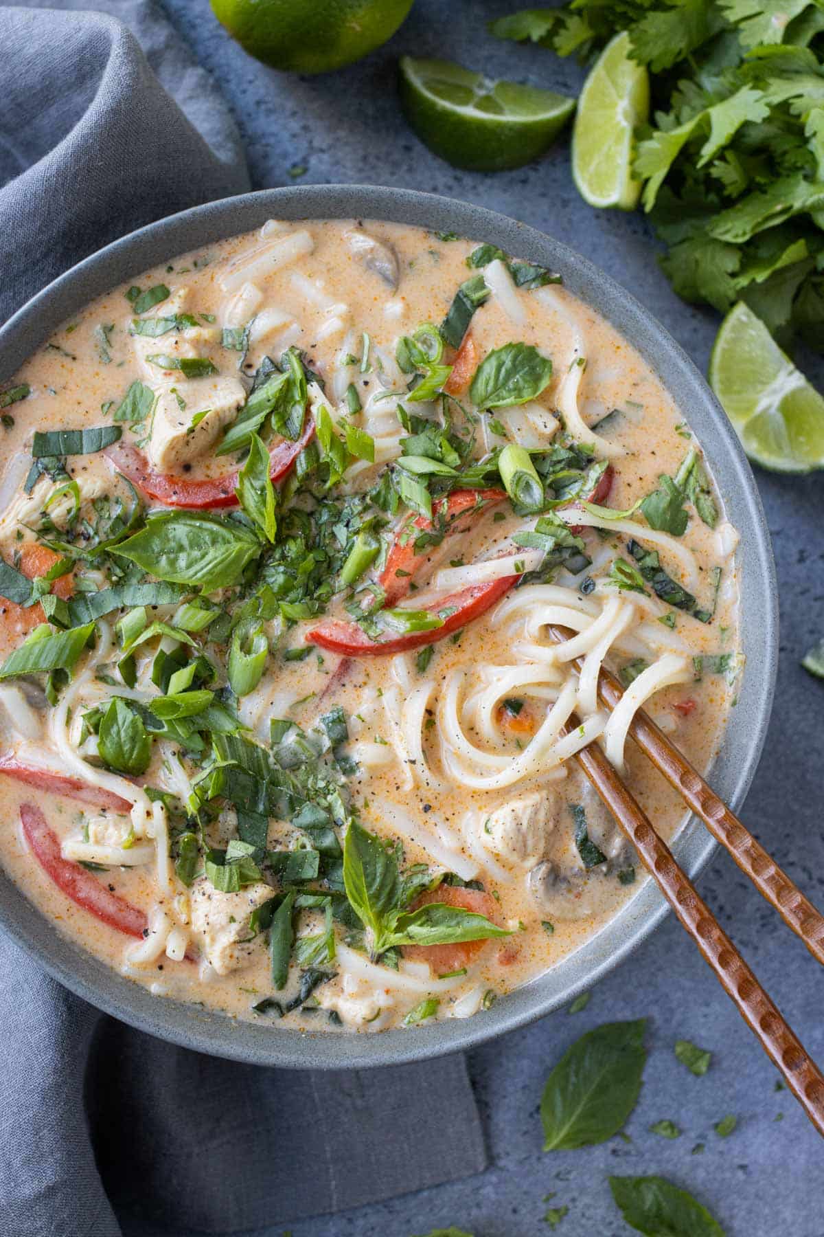 Chopsticks going into a bowl of Thai Red Curry Chicken Soup with fresh herbs and limes next to it.