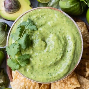 A bowl full of creamy avocado salsa verde is on a plate next to tortilla chips, avocados, and tomatillos.