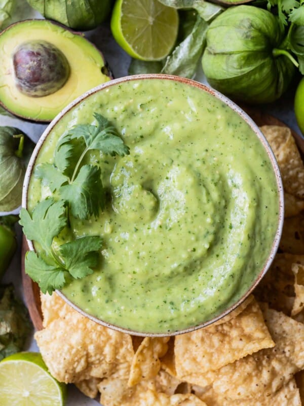 A bowl full of creamy avocado salsa verde is on a plate next to tortilla chips, avocados, and tomatillos.