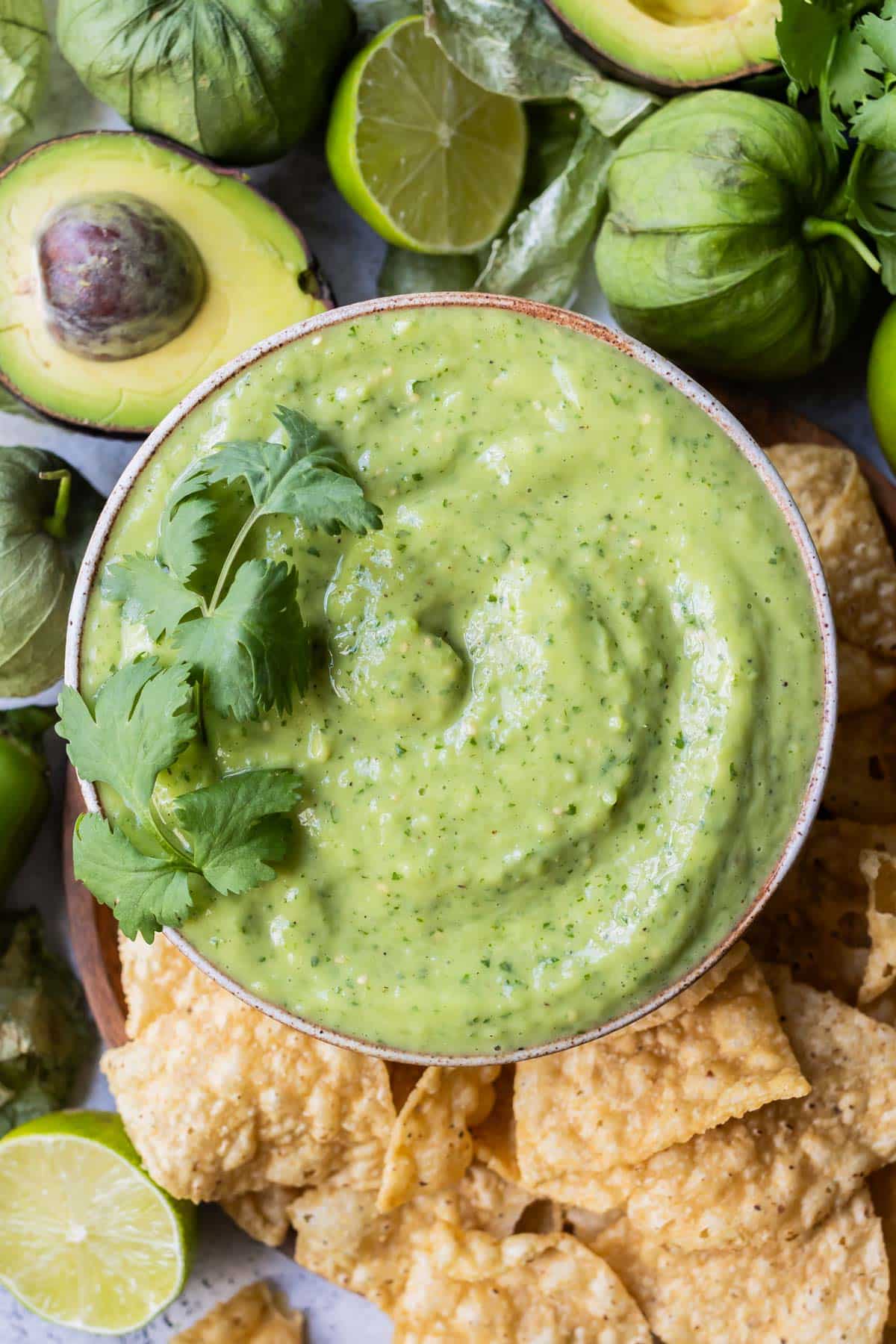 A bowl full of creamy avocado salsa verde is on a plate next to tortilla chips, avocados, and tomatillos.