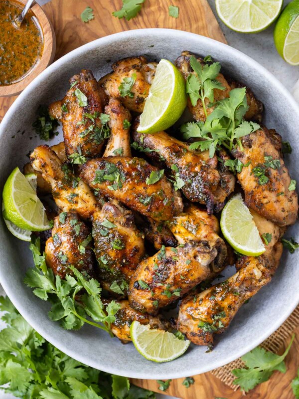 A bowl full of cilantro lime chicken wings that have been oven-baked sits on a wooden cutting board with cilantro sauce on the side.