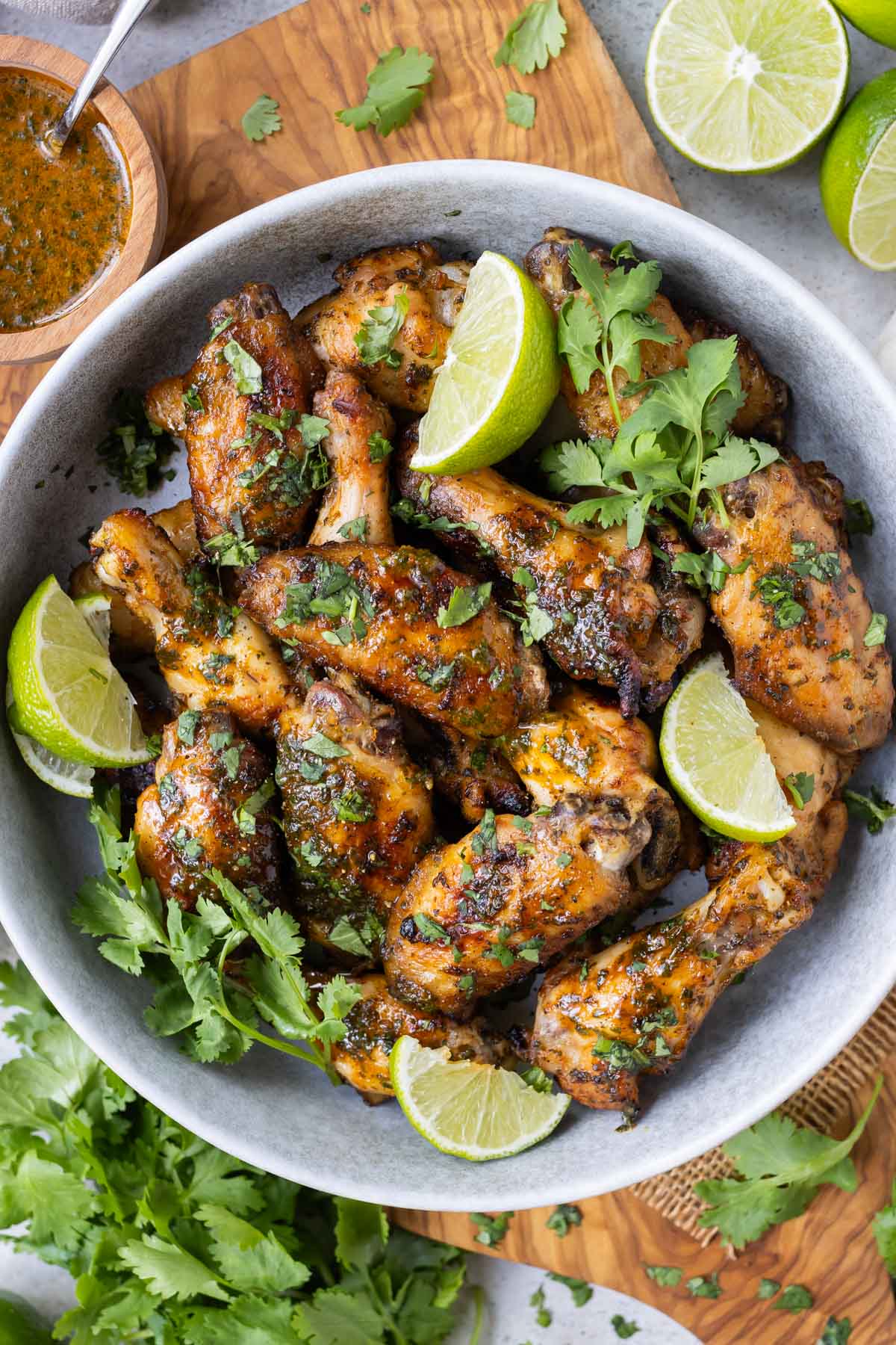 A bowl full of cilantro lime chicken wings that have been oven-baked sits on a wooden cutting board with cilantro sauce on the side.