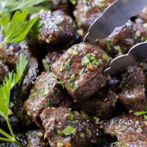 A garlic butter steak bite with fresh parsley being picked up with tongs from a cast iron skillet.