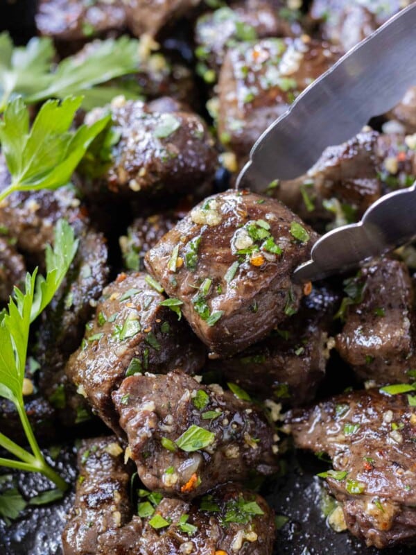 A garlic butter steak bite with fresh parsley being picked up with tongs from a cast iron skillet.