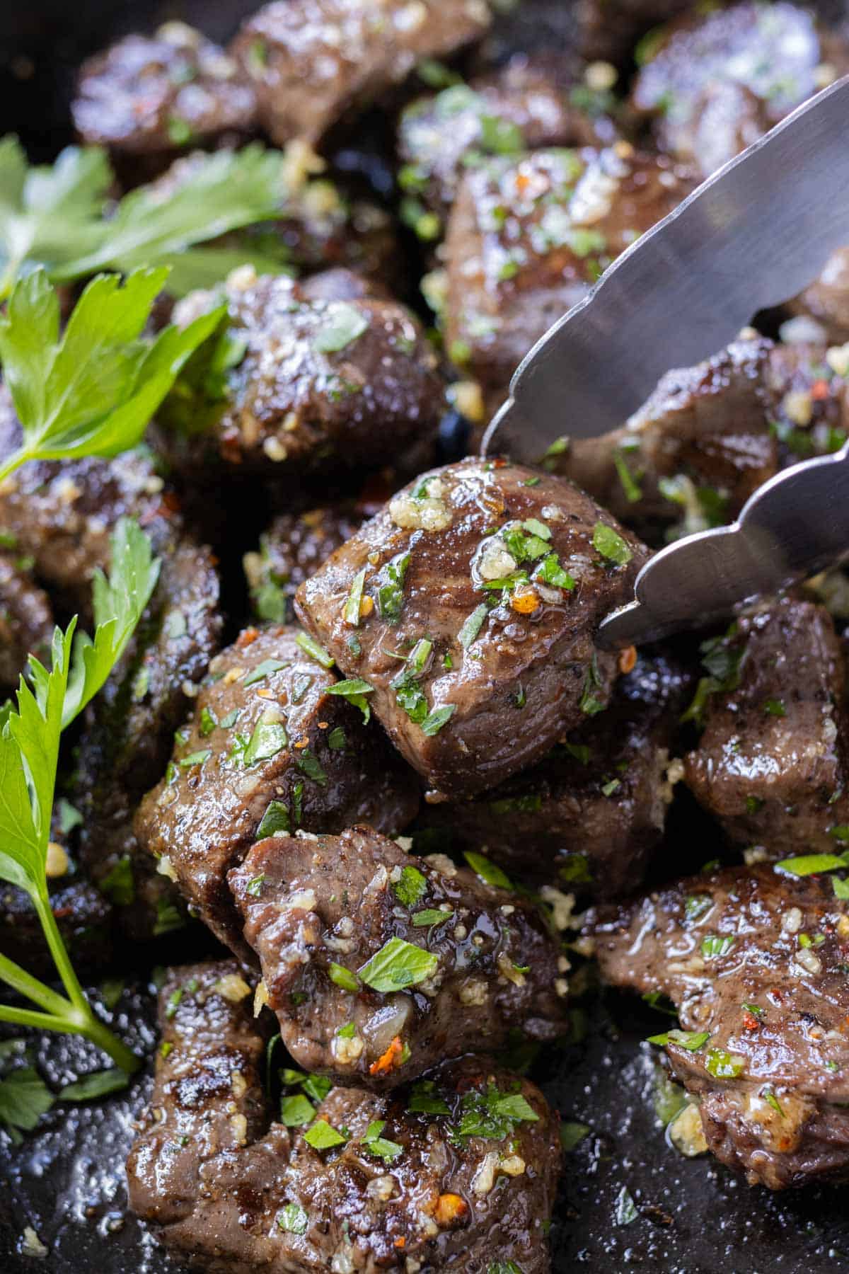 A garlic butter steak bite with fresh parsley being picked up with tongs from a cast iron skillet.