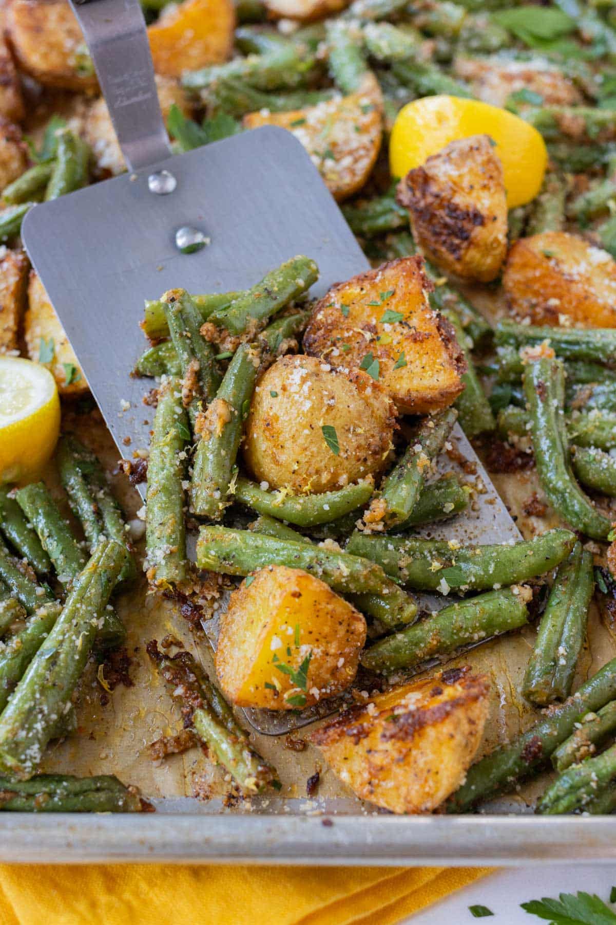 A spatula stirs up roasted potatoes and green beans topped with Parmesan and lemon.