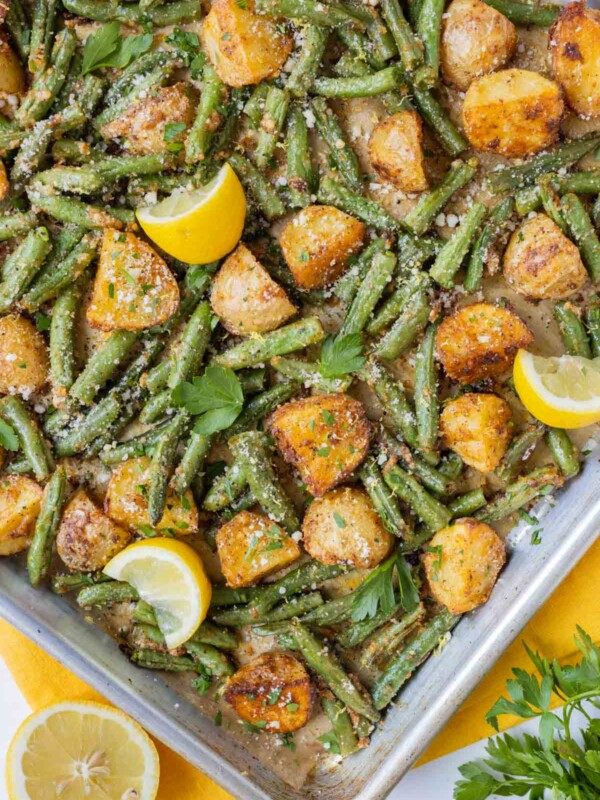 A sheet pan full of roasted potatoes and green beans with parmesan is next to a lemon and a yellow napkin.