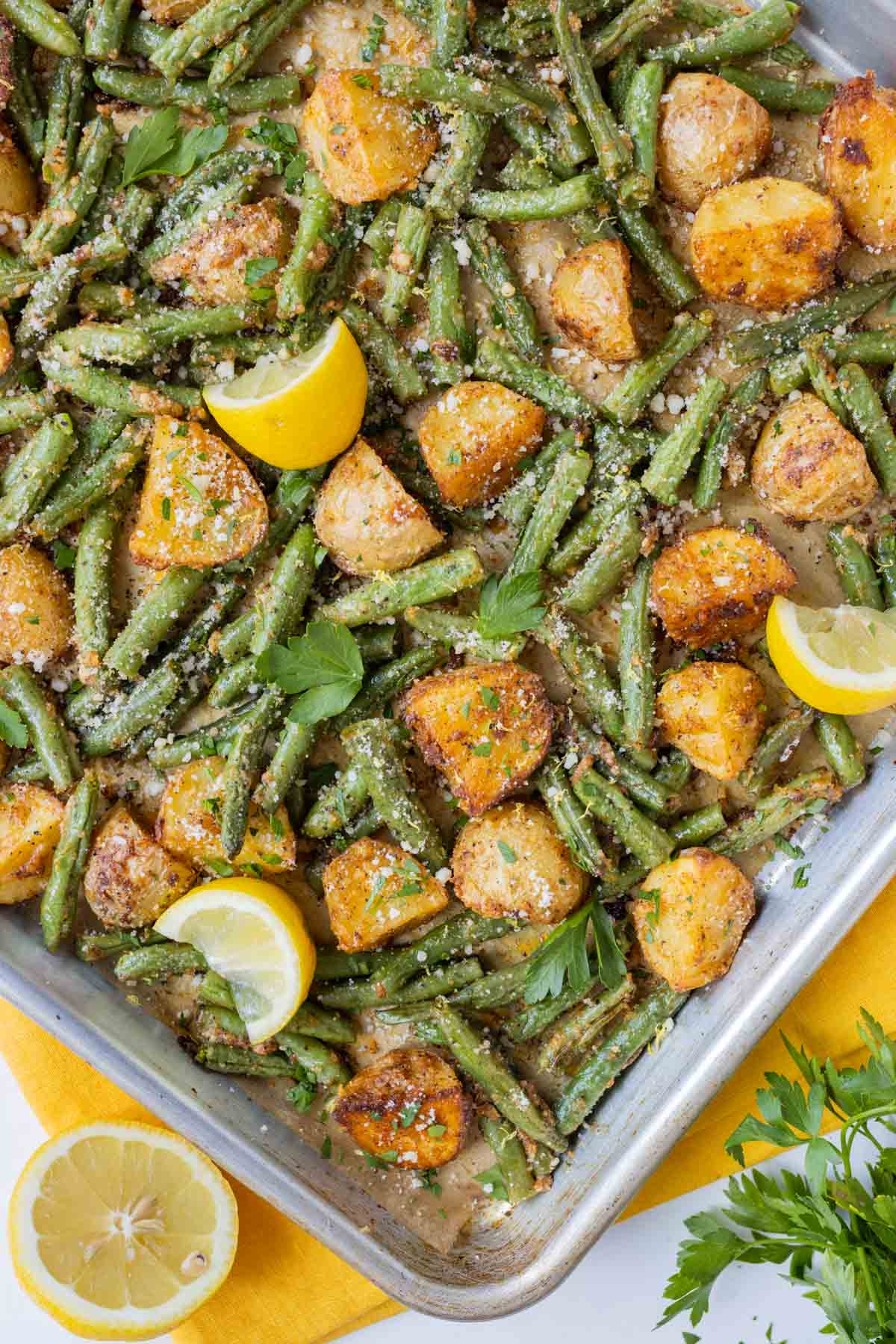 A sheet pan full of roasted potatoes and green beans with parmesan is next to a lemon and a yellow napkin.